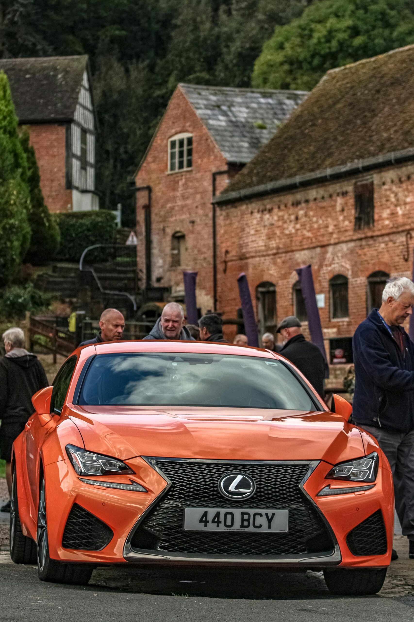 A sleek orange Lexus parked on a quaint street with historic buildings and people gathered around.